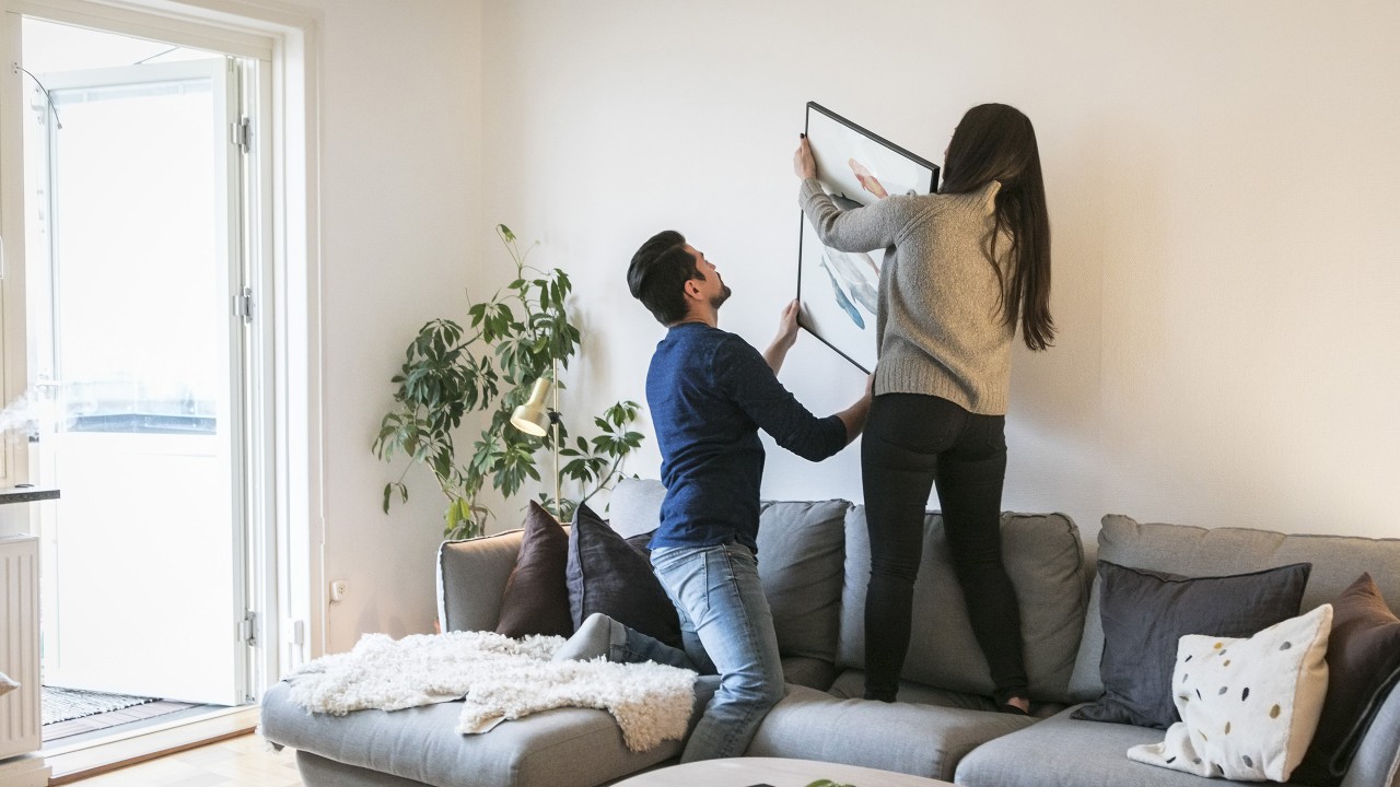 Couple standing on the couch to hang a picture on the wall; image used for Home Loan for NRI page.