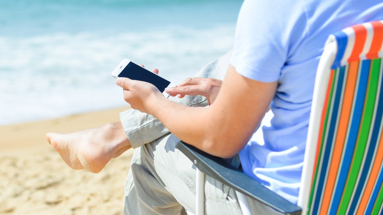 A man sitting on the beach with his mobile phone; image used for HSBC Mobile banking page.